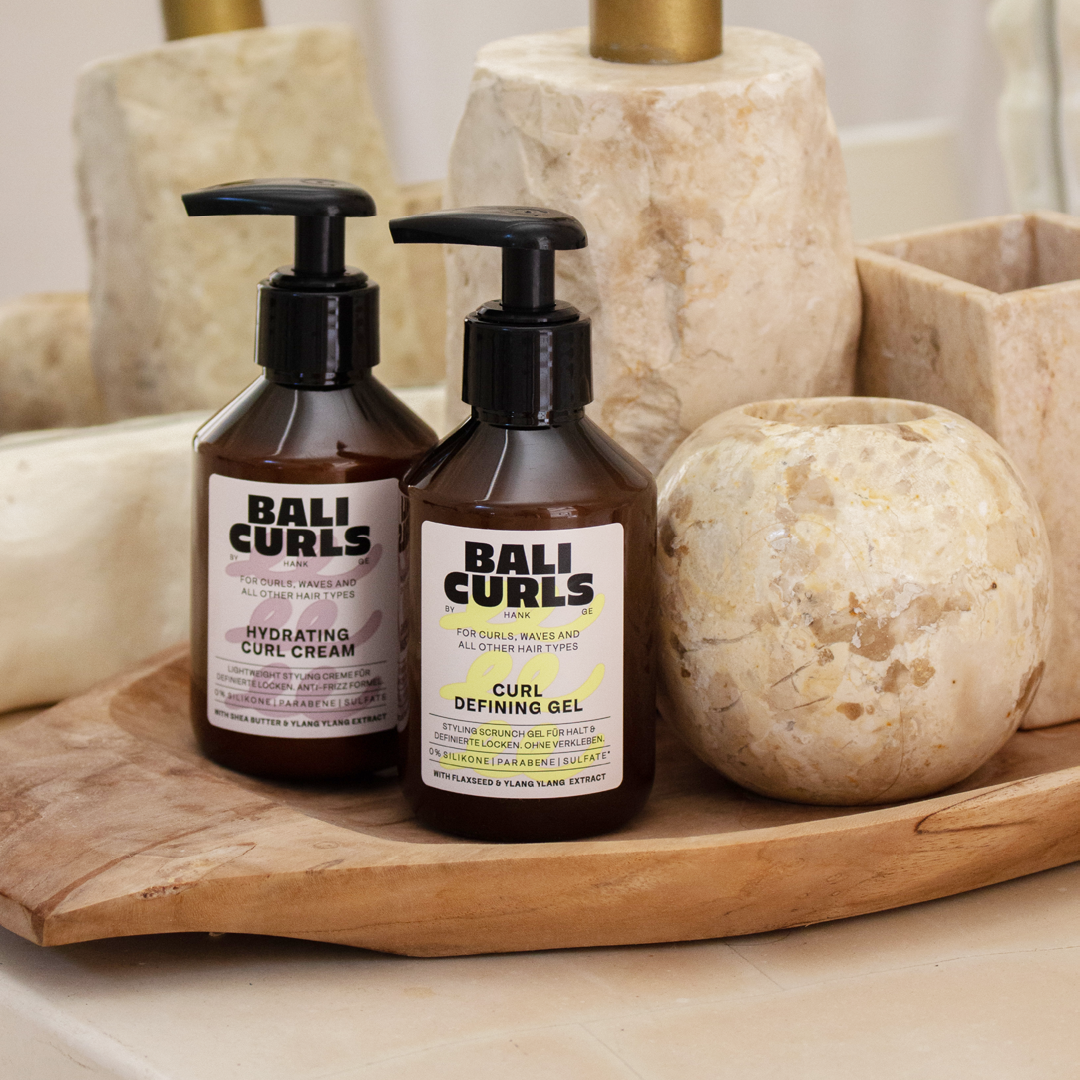 Abbildung des Bali Curls Curl Defining gels und der Hydrating Curl Cream auf einer Holzablage neben Steinernen Badezimmer Objekten